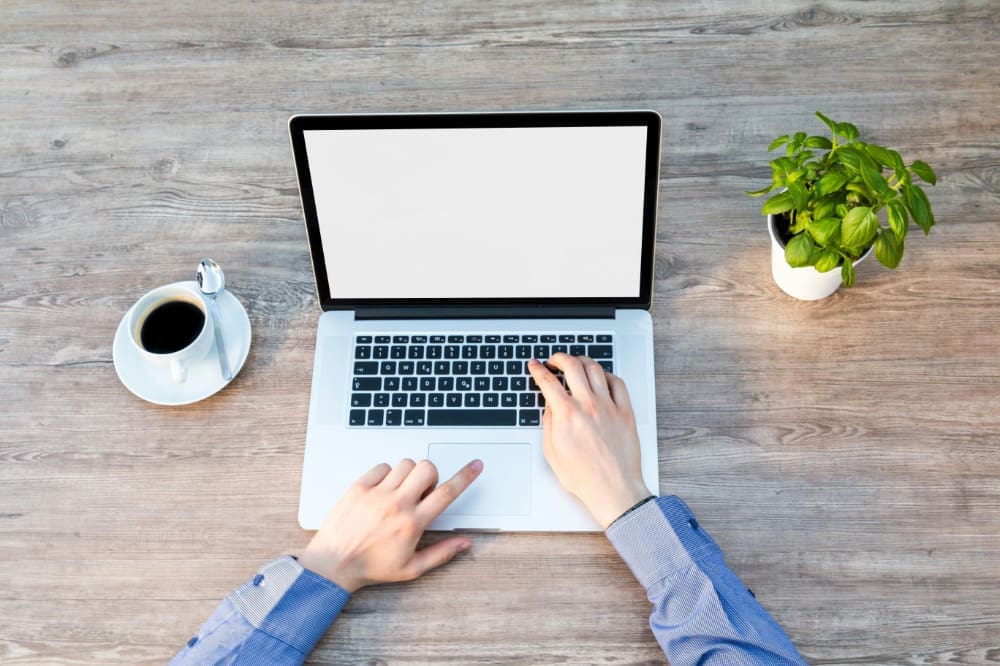 Freelancer-laptop Person typing on laptop rested on wooden desk outside, between a cup of coffee and a desk plant.