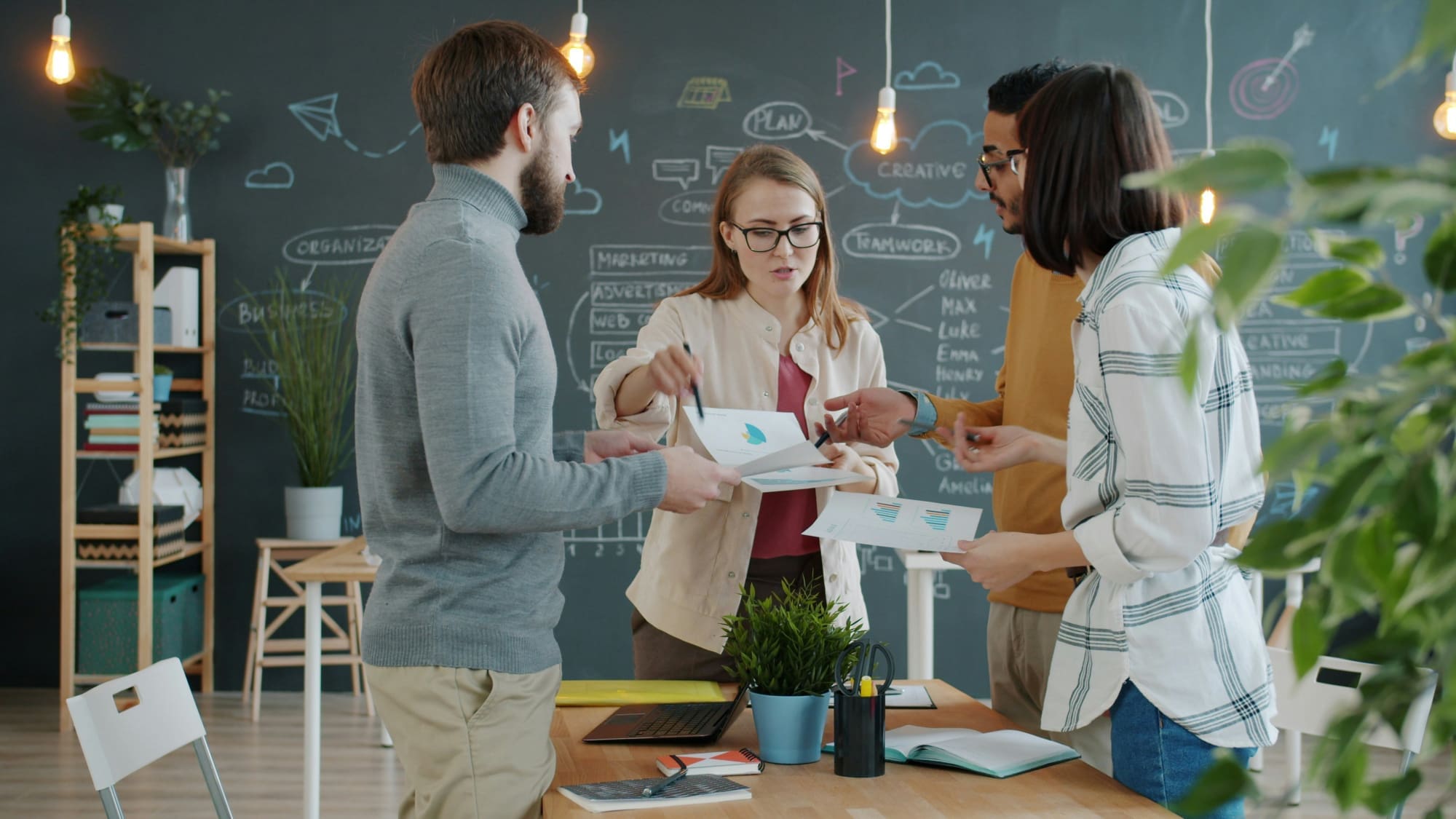 Four business people standing over desk discussing documents with blackboard in the background.
