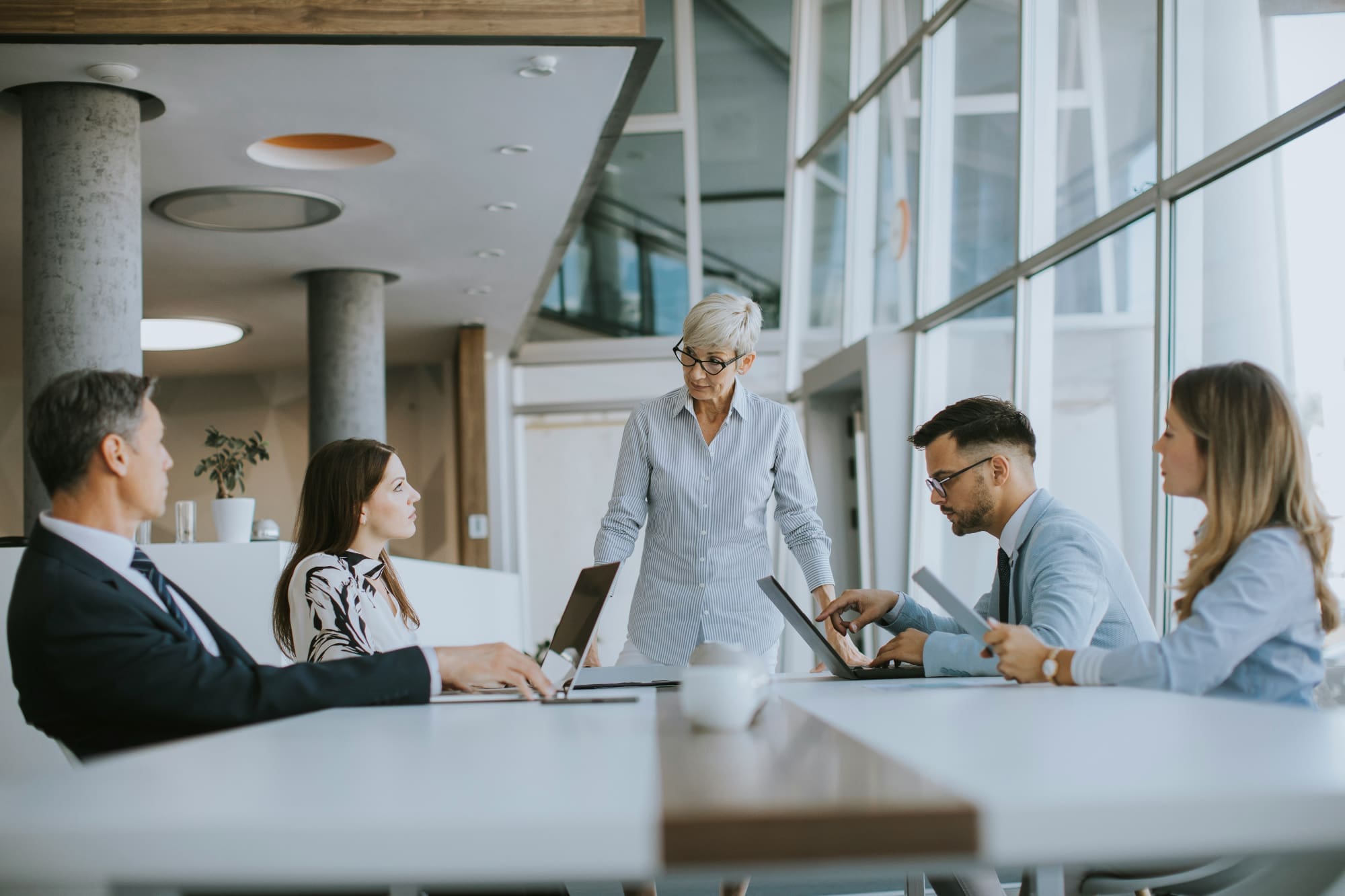 One business person stood at end of desk with others sitting down looking at her.